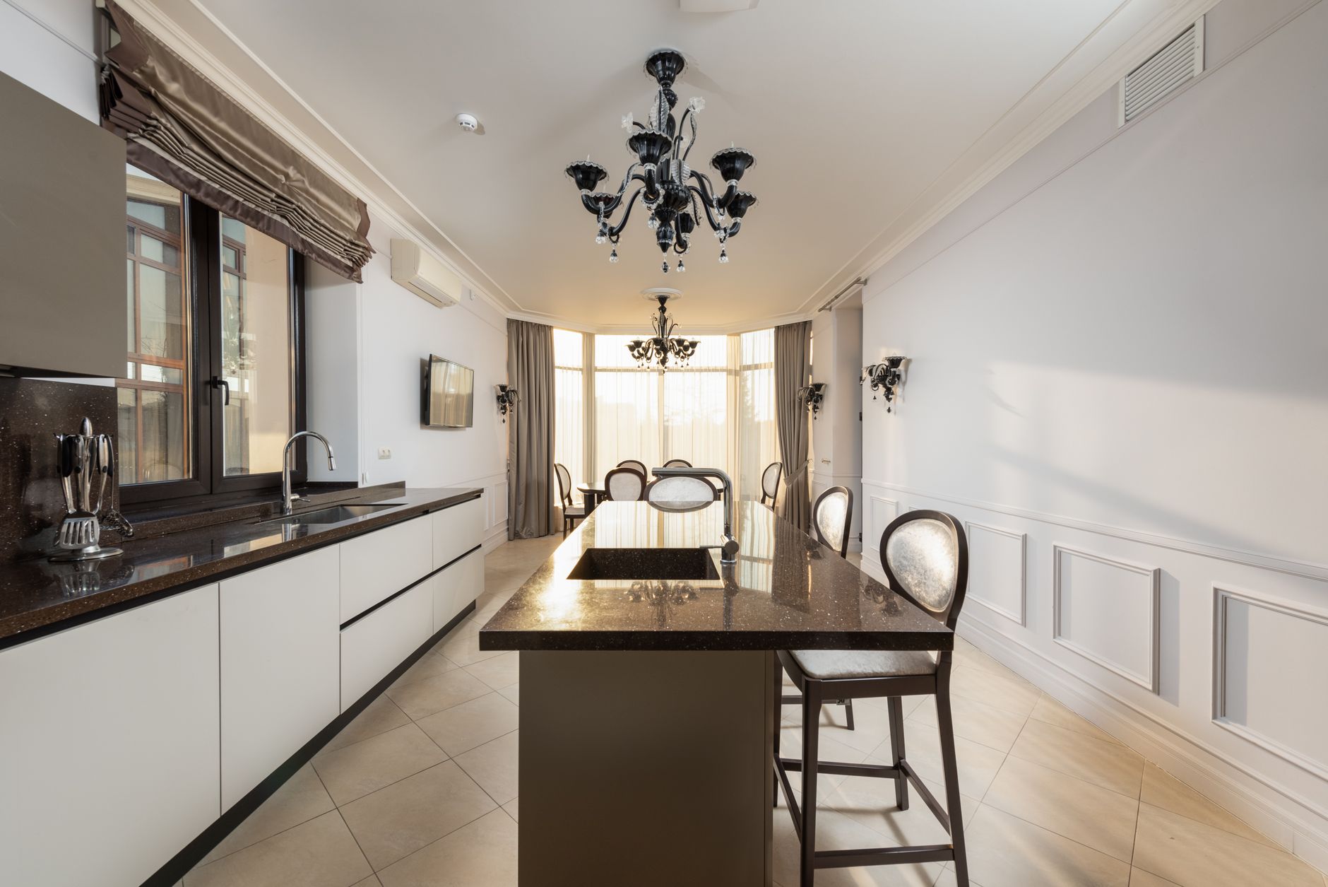 Chairs at table with sink near white cupboards and counter with supplies in spacious modern kitchen with window at home