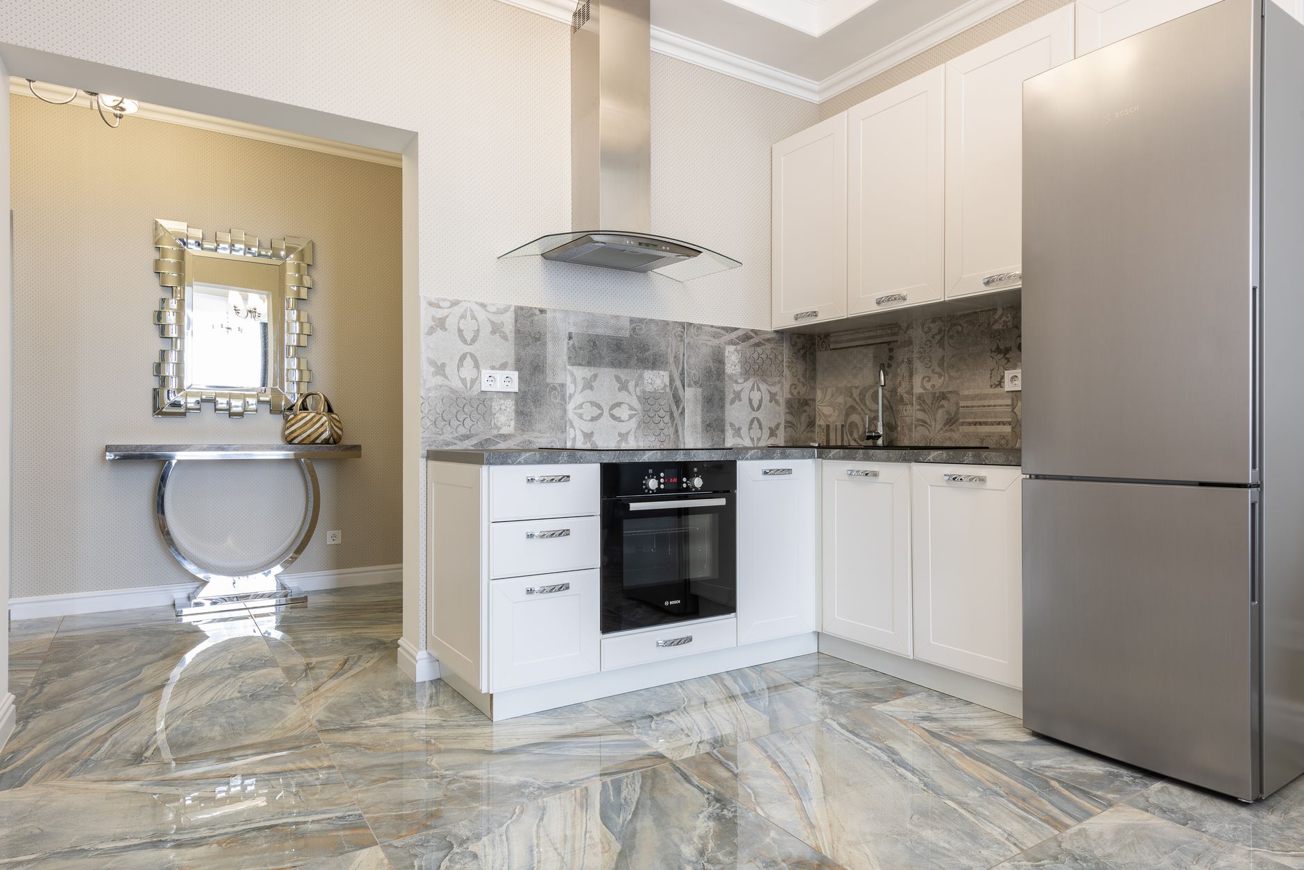 Contemporary kitchen with fridge and built in electric oven under hood in house with mirror and marble floor